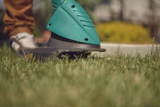 Guy in sneakers and pants is cutting green grass with professional electrical mini lawn mower on his backyard. Gardening care equipment and services. Sunny day. Close up