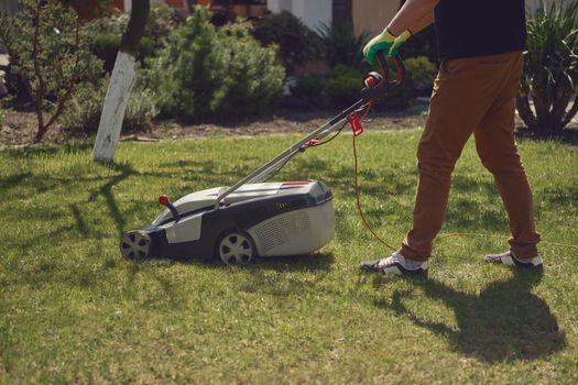 Strong male in casual outfit and colorful gloves is cutting green grass with electric lawn mower in his garden. Gardening care equipment and services. Sunny day