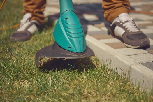 Male in sneakers and pants is cutting green grass with handheld modern lawn mower in his garden. Gardening care equipment and services. Sunny day. Close up