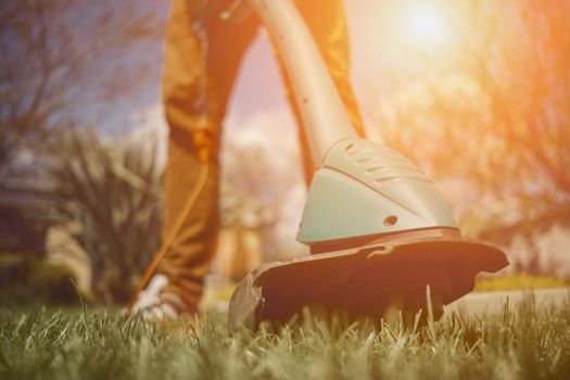 Male in casual outfit is mowing green grass with handheld electric lawn mower on his backyard. Gardening care tools and services. Sunny day. Close up