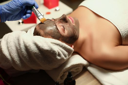 Top view of spa centre worker applying clay mask on clients face with special tool. Woman relaxing in cozy atmosphere. Wellness, skincare, beauty concept