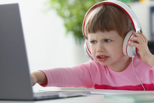 Portrait of little child not satisfied with something, point with finger on laptop screen. Upset kid in headset watch cartoon. Childhood, emotion concept
