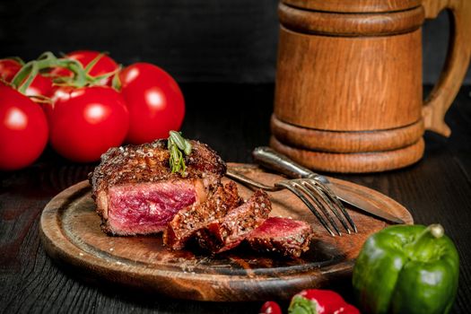 Grilled steak seasoned with spices and fresh herbs served on a wooden board with wooden mug of beer, fresh tomato, red and green peppers. Black wooden background. Still life