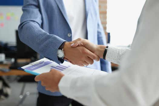 Close-up of partners shake hands over business statistics report paper. Biz partners perform friendly gesture after successful agreement. Contract concept