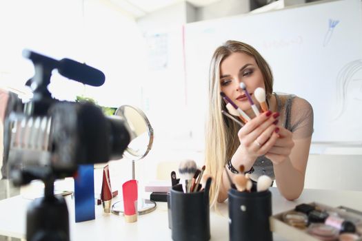 Portrait of pretty lady holding bunch of makeup brushes while recording video. Gorgeous woman making record for vlog on cosmetic product. Blogger concept