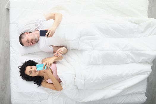 Top view of wife and husband laying in bed staring at phones screens from early morning. Wake up and fall asleep with smartphone. Gadget addiction concept