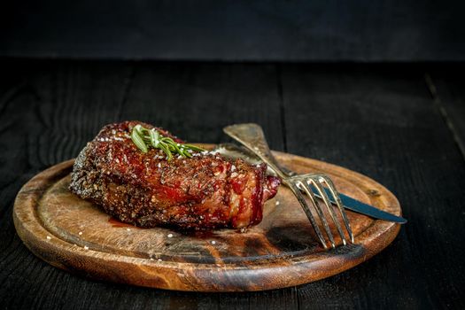 Barbecue Dry aged Ribeye Steak with knife and fork on cutting board. Black wooden background. Still life. Copy space. Close-up