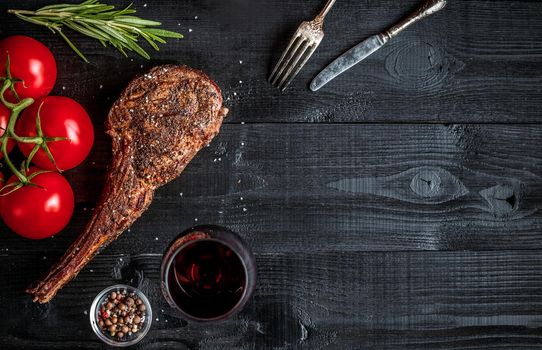 Barbecue dry aged rib of beef with spice, vegetables and glass of red wine close-up on black wooden background. Top view. Copy space. Still life. Flat lay