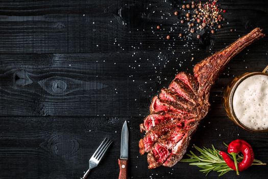 Barbecue dry aged rib of beef with spice, vegetables and a glass of light beer close-up on black wooden background. Top view. Copy space. Still life. Flat lay