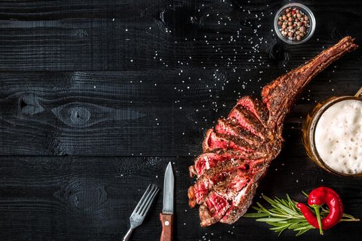 Barbecue dry aged rib of beef with spice, vegetables and a glass of light beer close-up on black wooden background. Top view. Copy space. Still life. Flat lay