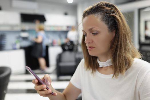 Portrait of woman client sit at hairdresser, woman use smartphone while waiting for hair to dye. Female enjoy beauty procedure. Wellness, self care concept