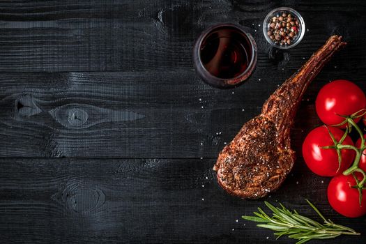 Barbecue dry aged rib of beef with spice, vegetables and glass of red wine close-up on black wooden background. Top view. Copy space. Still life. Flat lay