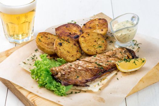 Fish fillet, fried potatoes with greens, lemon and sauce on a wooden board. A glass of light beer and a ready fish dish on a white wooden table