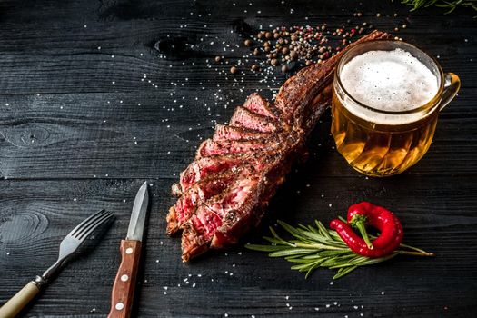 Barbecue dry aged rib of beef with spice, vegetables and a glass of light beer close-up on black wooden background. Top view. Copy space. Still life. Flat lay