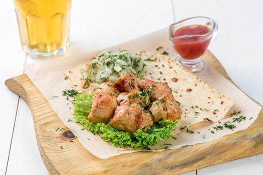 Main course of chicken brisket pieces served with greens and sauce on a wooden board. A glass of light beer and chicken brisket on a white wooden table