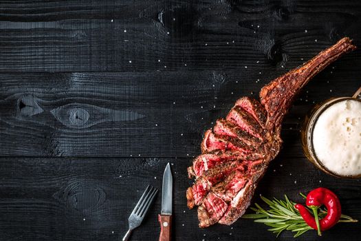 Barbecue dry aged rib of beef with spice, vegetables and a glass of light beer close-up on black wooden background. Top view. Copy space. Still life. Flat lay