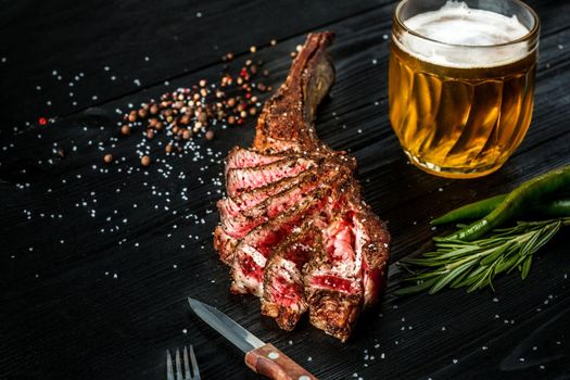 Barbecue dry aged rib of beef with spice, vegetables and a glass of light beer close-up on black wooden background. Top view. Copy space. Still life. Flat lay