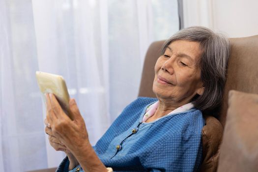 Elderly asian woman relax on couch in living room browsing wireless Internet on tablet