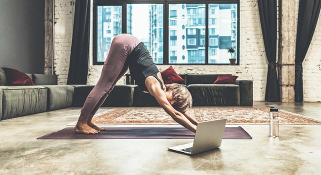 Nice Woman Practicing Yoga, Doing Down Dog Exercise, Watching Online Yoga Lessons by her Laptop, Yoga at Home. Yoga Time, Woman Relaxing in her Apartment in Megapolis City. Multi-storey Residential Building Background