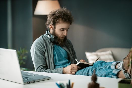 Student Listens to an Online Lecture and Makes Notes in a Notebook. a Man Writes Important Information in a Diary. Headphones Around his Neck. Feet on the Table. Close-up. High quality photo