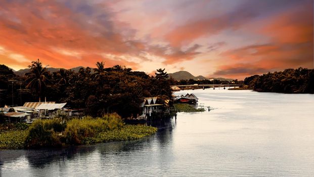 Dark contrast sunset in tropics on big river with village, bridge and mountains on background. The Bridge on the River Kwai, Thailand 5th of july 2009. High quality photo