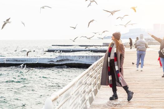 Walking woman on the pier in Winter time. Stylish lady wearing winter coat, rough boots, scarf and hat standing alone near the sea
