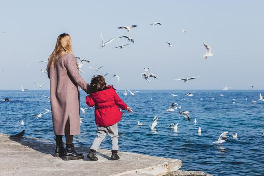 Mother and son walk to the seaside. Birds flying above the water. Cold season vacations