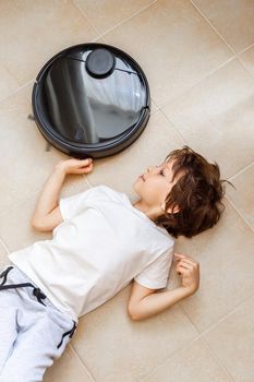 Kid playing with Robotic Vacuum Cleaner. Boy lying on the floor in the way of the Vacuum Cleaner route