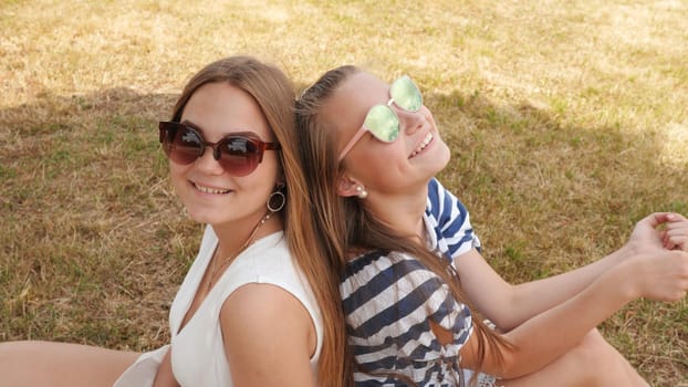 Cheerful female friends in sunglasses are sitting on the grass in the city