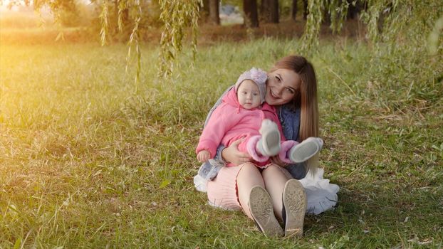 Happy young mother with her baby is sitting on the grass in the park in the autumn evening