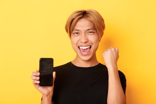 Close-up of happy rejoicing asian guy showing smartphone screen and saying yes, fist pump as triumphing, winning or achieve goal, yellow background.