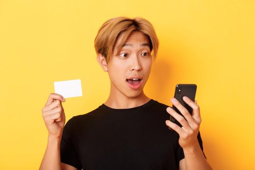 Close-up of amazed asian guy looking at mobile phone while shopping online and showing credit card, yellow background.