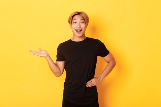 Portrait of happy and excited smiling asian guy holding something on hand over yellow background.