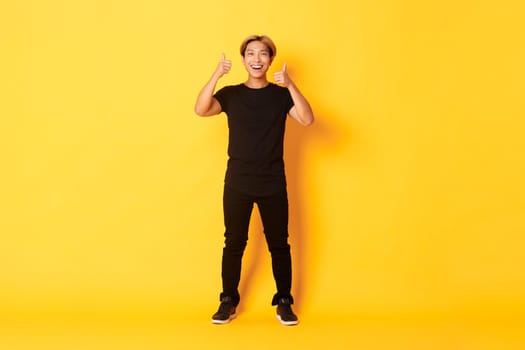 Full-length of satisfied handsome asian guy smiling, showing thumbs-up in approval, yellow background.