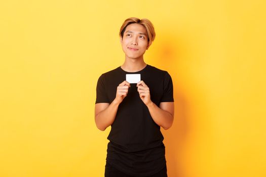 Thoughtful smiling asian guy thinking while showing credit card, looking upper left corner dreamy, yellow background.