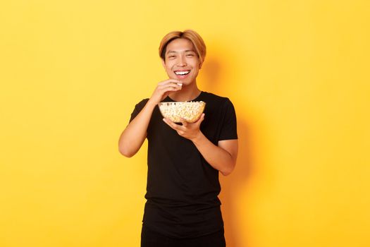 Carefree happy asian guy with blond hair, watching comedy, laughing and eating popcorn, standing yellow background.