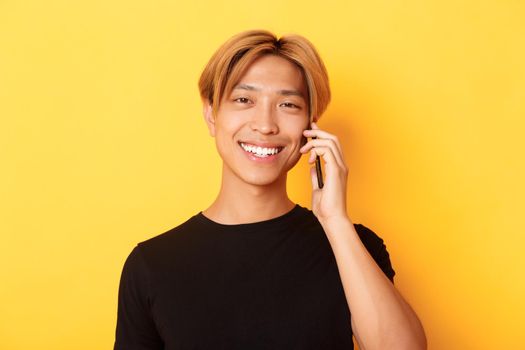 Close-up of handsome stylish korean guy smiling and talking on mobile phone, standing over yellow background.