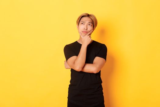 Portrait of thoughtful attractive guy in black t-shirt looking upper left corner. Young man pondering idea, standing yellow background.