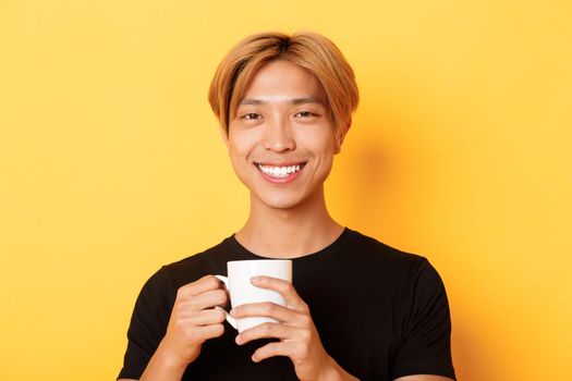 Close-up of happy satisfied asian smiling guy, holding mug with coffee, drinking and looking pleased, standing over yellow background.