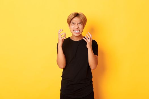 Portrait of angry and pissed-off asian blond guy, clenching hands furious and grimacing, standing over yellow background.