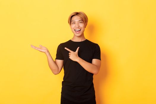 Portrait of handsome smiling asian guy holding something on hand and pointing finger at it, standing yellow background pleased.