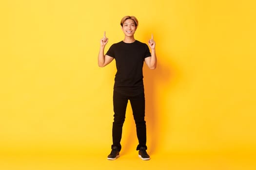 Full-length of confident handsome asian man with blond hair, smiling and pointing fingers up, showing logo, yellow background.