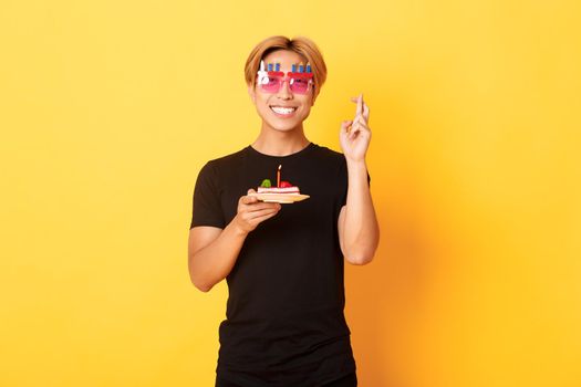 Portrait of hopeful attractive asian birthday guy, celebrating in funny party glasses, holding cake and fingers crossed while making b-day wish, yellow background.