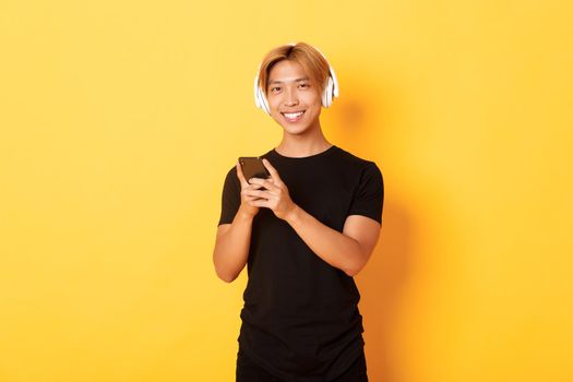 Good-looking smiling, pleased asian guy in wireless headphones, holding smartphone and looking satisfied camera, listening music or podcast.