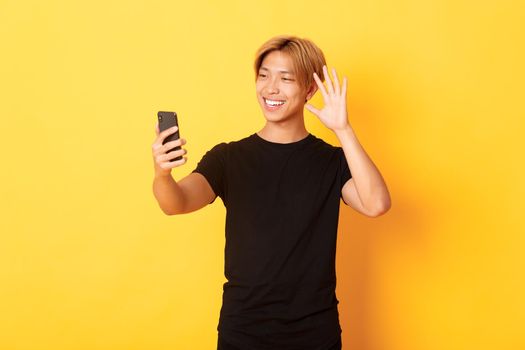 Portrait of friendly handsome asian man, smiling and waving hand at smartphone camera, greeting friends on video call, standing yellow background.