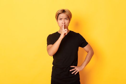 Portrait of angry asian man frowning, shushing at camera disappointed, standing yellow background.