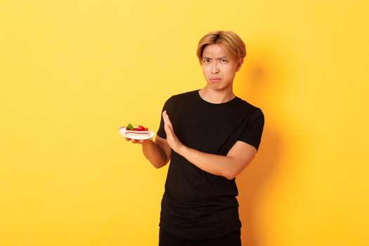 Portrait of disgusted and upset young asian guy refuse eating cake, looking displeased and showing rejection gesture, yellow background.