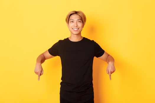 Portrait of smiling attractive korean guy with blond hair, wearing black outfit, pointing fingers down, showing way, standing yellow background.