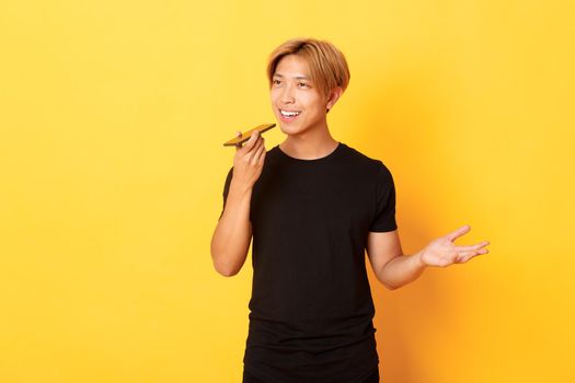 Portrait of stylish handsome korean guy with blond hair record voice message on mobile phone, holding smartphone near mouth and speaking, standing yellow background.