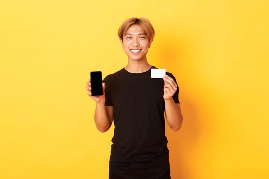 Portrait of handsome young asian guy showing smartphone screen, banking app and credit card, standing yellow background and smiling.